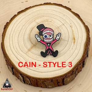 Puede incluir: Un pin de dibujos animados con un personaje con un traje rosa, sombrero de copa negro y boca abierta con dientes. El pin tiene el texto "CAIN - STYLE 3" en rojo. El pin est&aacute; sobre una rodaja de madera.
