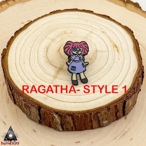 Puede incluir: Un charm de Ragatha-Style 1, que presenta un personaje de dibujos animados con cabello rosado, un ojo azul y un vestido morado. El charm est&aacute; colocado sobre una rodaja de madera con el texto "RAGATHA- STYLE 1" en rojo.