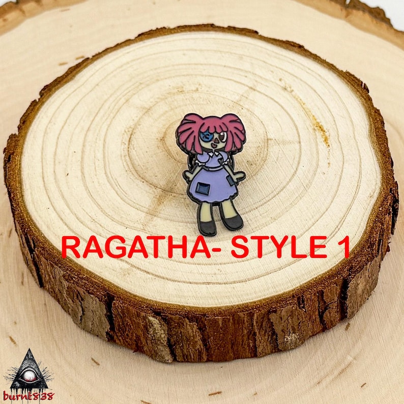 Puede incluir: Un charm de Ragatha-Style 1, que presenta un personaje de dibujos animados con cabello rosado, un ojo azul y un vestido morado. El charm est&aacute; colocado sobre una rodaja de madera con el texto "RAGATHA- STYLE 1" en rojo.