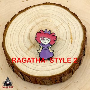 Puede incluir: Un colorido pin de esmalte con una mu&ntilde;eca de dibujos animados con pelo rosa, un vestido morado y un ojo gui&ntilde;ando. El pin est&aacute; colocado sobre una rodaja de madera, con el texto "RAGATHA - STYLE 2" en rojo.