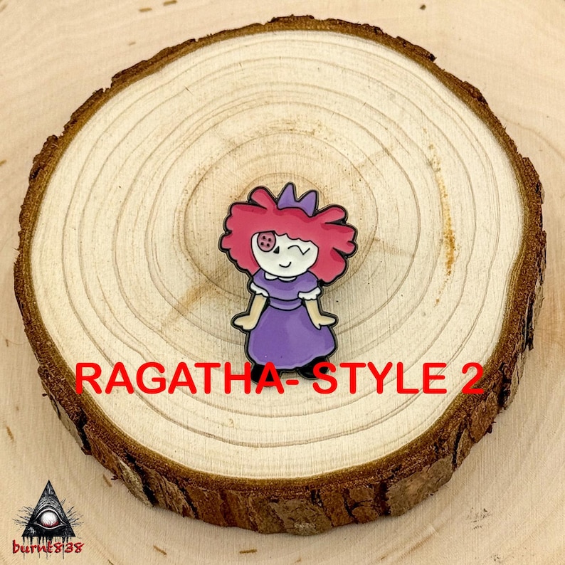 Puede incluir: Un colorido pin de esmalte con una mu&ntilde;eca de dibujos animados con pelo rosa, un vestido morado y un ojo gui&ntilde;ando. El pin est&aacute; colocado sobre una rodaja de madera, con el texto "RAGATHA - STYLE 2" en rojo.