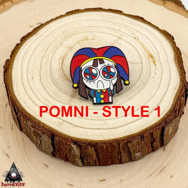 Puede incluir: Un pin de dibujos animados colorido con un personaje triste con ojos grandes y detallados y un sombrero de buf&oacute;n rojo, azul y amarillo. El pin est&aacute; etiquetado como "POMNI - STYLE 1" en texto rojo. Sobre una rodaja de madera.