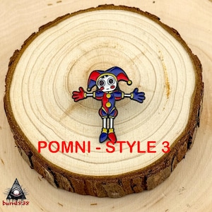 Puede incluir: Un pin de esmalte colorido con un dise&ntilde;o de buf&oacute;n con acentos rojos, azules y amarillos. El pin tiene los brazos extendidos y est&aacute; sobre un fondo de madera. Se muestra el texto "POMNI - STYLE 3".