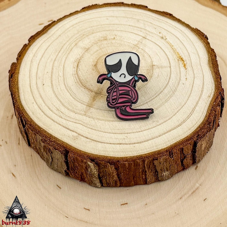Puede incluir: Un pin de esmalte negro y rosa con un personaje de dibujos animados triste con una cabeza blanca, ojos negros y l&aacute;grimas rosas. El personaje est&aacute; envuelto en espirales rosas. El pin est&aacute; colocado sobre una rodaja de madera.
