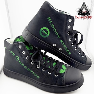 May include: Black high-top sneakers with green text that reads "TYPE O NEGATIVE" and "BLOODY KISSES". The sneakers have black laces, a black sole, and a green logo. The shoes are made of canvas.