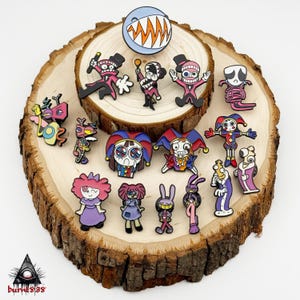 Puede incluir: Una colecci&oacute;n de coloridos pines de esmalte con varios personajes de dibujos animados con dise&ntilde;os &uacute;nicos. Los pines incluyen personajes con sombreros de copa, sombreros de buf&oacute;n y otros elementos caprichosos. Los pines se exhiben sobre una rodaja de madera.