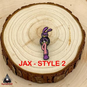 Puede incluir: Un pin de conejo de dibujos animados p&uacute;rpura con una sonrisa traviesa, que lleva pantalones rosas y sostiene un objeto negro. El pin est&aacute; etiquetado "JAX - STYLE 2" en texto rojo. El pin est&aacute; colocado sobre una rodaja de madera.