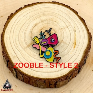 Puede incluir: Un colorido pin de esmalte con un personaje de dibujos animados con cabeza rosa, cuerpo amarillo y ojo morado. El pin tiene el texto "ZOOBLE - STYLE 2" en rojo. El pin est&aacute; sobre una rodaja de madera.