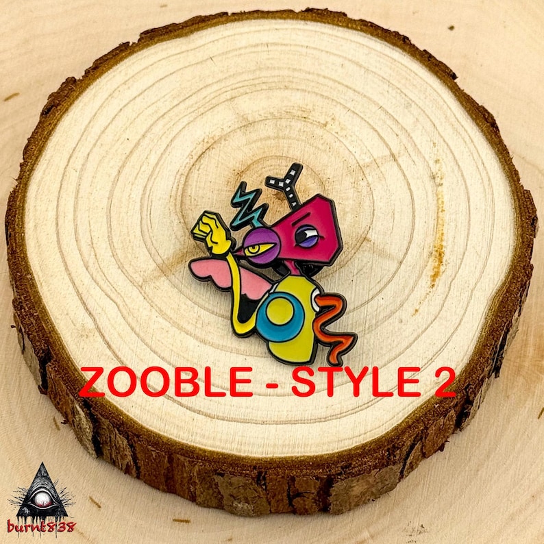 Puede incluir: Un colorido pin de esmalte con un personaje de dibujos animados con cabeza rosa, cuerpo amarillo y ojo morado. El pin tiene el texto "ZOOBLE - STYLE 2" en rojo. El pin est&aacute; sobre una rodaja de madera.