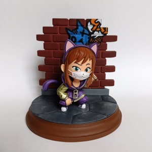 May include: A small figurine of a girl wearing a purple hoodie with cat ears and a white face mask. She is holding a stick and standing in front of a brick wall with graffiti.
