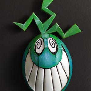 Puede incluir: Una máscara de cerámica verde y azul con una amplia sonrisa de dientes y ojos en espiral. La máscara tiene una cresta verde y dentada en la parte superior.