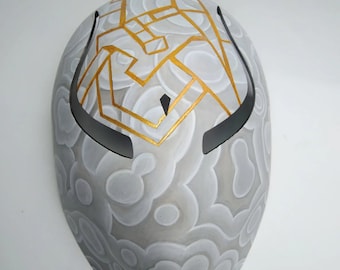 Op de afbeelding: Een wit masker met een geometrisch patroon in gouden verf. Het masker heeft een afgeronde vorm met twee ooggaten en een puntige kin.