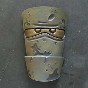 Puede incluir: Una maceta decorativa con temática ninja verde y dorada. La maceta tiene una superficie texturizada con un diseño de máscara ninja. Los ojos y los detalles de la máscara son dorados, y el resto de la maceta es verde.