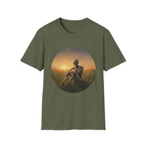 May include: Olive green t-shirt featuring a circular graphic of a figure sitting in a field at sunset. The figure is pale with a wooden appearance. The background shows a golden sunset with a field of grass and flowers.