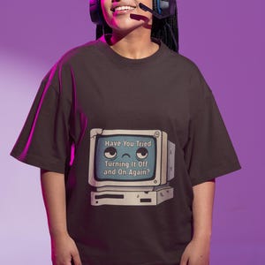 May include: A dark brown short-sleeved t-shirt featuring a graphic of a vintage computer monitor with a sad face and the text "Have You Tried Turning It Off and On Again?" printed on the front. The shirt has a crew neckline and a relaxed fit.