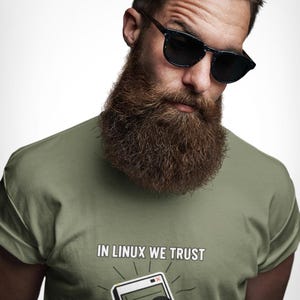 May include: An olive green t-shirt featuring a graphic of a penguin wearing a red bowtie next to a computer window icon. Above the graphic, the text reads IN LINUX WE TRUST in white capital letters. The shirt has short sleeves and a crew neckline.