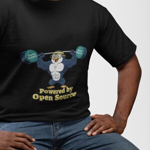 May include: A black short-sleeve t-shirt featuring a graphic of a muscular penguin wearing glasses and a headband, lifting a barbell with weights labelled Open Source. Below the penguin, the text reads Powered by Open Source in yellow.