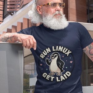 May include: A navy blue short-sleeved t-shirt featuring a circular graphic with a penguin wearing sunglasses. The text RUN LINUX GET LAID is printed in a circular arc around the penguin, accented by small colorful hearts and star shapes.