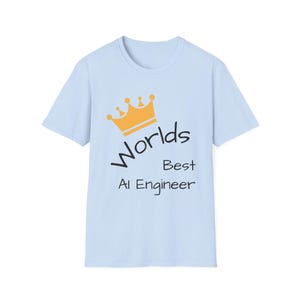 May include: A pale blue t-shirt featuring the text "Worlds Best AI Engineer" in black, with a gold crown graphic above. The shirt has a crew neck and is likely made of a soft fabric.