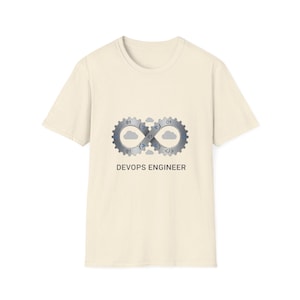 Puede incluir: Camiseta color crema con un gráfico de dos engranajes entrelazados con símbolos de nube y código, formando un símbolo de infinito. El texto "DEVOPS ENGINEER" está debajo. La camiseta está hecha de una tela suave y texturizada.