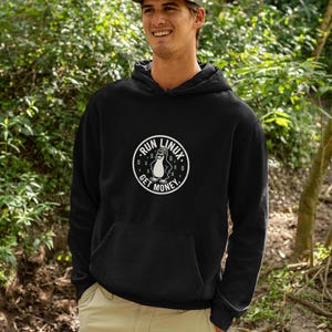 Pode incluir: Uma camisola preta com capuz e um gráfico circular no peito. O gráfico mostra um pinguim com um gorro, rodeado pelo texto RUN LINUX GET MONEY e sinais de dólar. A camisola tem mangas compridas e um bolso frontal tipo canguru.