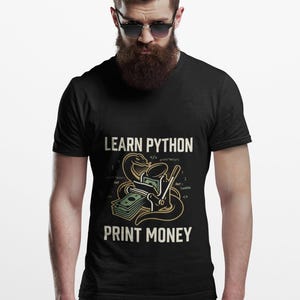 May include: A black short-sleeved t-shirt featuring a graphic print with the text LEARN PYTHON and PRINT MONEY. The design includes a snake coiled around a printing press that is producing stacks of green paper currency, along with code snippets.