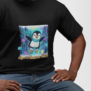May include: A black t-shirt featuring a graphic of a cartoon penguin wearing a light blue scarf and a utility belt. The penguin is positioned in front of a stylised city background with neon accents. The text Open Source Hero is printed below.