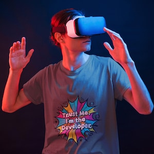 May include: A grey short-sleeved t-shirt featuring a colorful comic-style graphic burst with the text "Trust Me, I'm the Developer" surrounded by small coding bracket symbols. The shirt is worn by an individual wearing a white and black VR headset.
