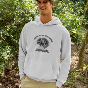 Puede incluir: Una sudadera con capucha de color gris claro que presenta un gráfico negro de un cerebro humano conectado a un patrón de circuito sobre una multitud de personas, con el texto I, FOR ONE, WELCOME OUR AI OVERLORDS impreso en arco.