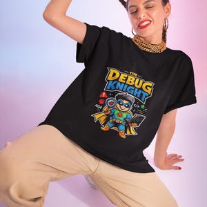 Puede incluir: Una camiseta negra de manga corta con un estampado gráfico de un personaje de superhéroe con capa y máscara, que sostiene un portátil y una lupa. El texto "THE DEBUG KNIGHT" está impreso sobre el personaje en letras amarillas y azules.