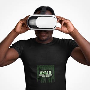 Op de afbeelding: Een zwart t-shirt met een groene digitale matrix-stijl afbeelding met de tekst "WHAT IF WE'RE IN A SIMULATION RIGHT NOW?" en een witte virtual reality-headset met een zwarte band en voorpaneel, gedragen over de ogen en het hoofd.