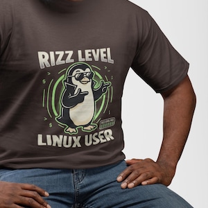 Pode incluir: Uma t-shirt de manga curta castanho-escura com um gráfico de um pinguim a usar óculos de sol e a apontar. O texto diz RIZZ LEVEL LINUX USER com uma barra de progresso RIZZ LEVEL MAX. O design inclui detalhes circulares verdes e símbolos.