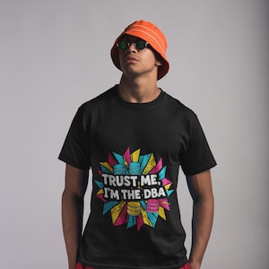 Könnte beinhalten: Ein schwarzes Kurzarm-T-Shirt mit einem Grafikdruck, der den Text "TRUST ME, I'M THE DBA" in weißen Buchstaben zeigt. Das Design enthält bunte geometrische Formen in Pink, Gelb und Blau sowie Illustrationen von gestapelten Münzen.