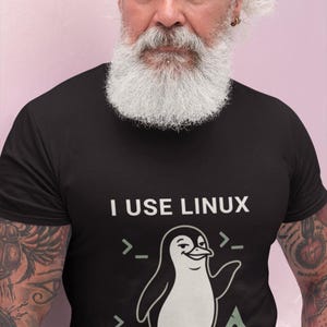 May include: A black short-sleeved t-shirt featuring a graphic of a penguin with a smug expression. The text reads I USE LINUX above the penguin and BY THE WAY below it. There are command line prompt symbols and an Arch Linux logo surrounding the penguin.