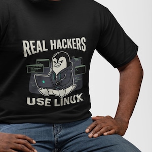 Pode incluir: Uma t-shirt preta com um gráfico de um pinguim de desenho animado a usar um capuz e a digitar num portátil. O texto acima do pinguim diz REAL HACKERS e o texto abaixo diz USE LINUX. O design inclui várias janelas de terminal de computador.