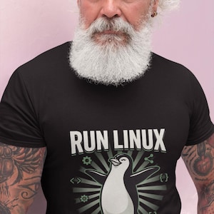 Puede incluir: Una camiseta negra de manga corta con un gráfico de un pingüino con las alas extendidas. Sobre el pingüino aparece el texto "RUN LINUX" y debajo "LIVE FREE". El diseño incluye iconos verdes de engranajes, corchetes y compartir.