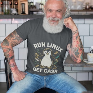 Pode incluir: Uma t-shirt cinzenta escura de manga curta com um gráfico de um pinguim com os braços estendidos, rodeado por notas flutuantes e código binário. O texto "RUN LINUX" está impresso acima do pinguim e "GET CASH" está impresso abaixo.
