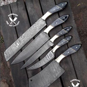 以下が含まれることがあります： 手作りの高級アートカトラリーナイフの詰め合わせ。刃には模様があり、黒い柄が付いています。ナイフは濃い木製の表面に配置されています。ハンドルには金色のボルスターと黒いエンドキャップが付いています。「HANDMADE FINE ART CUTLERY」のロゴが見えます。