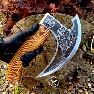 Peut inclure: Une hache à main avec une lame en métal incurvée présentant un motif de crâne gravé, un symbole circulaire et des runes. Le manche est en bois brun clair avec un grain visible. La lame en métal a un bord poli et une finition sombre.