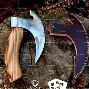 May include: A curved steel axe head with a wooden handle and a matching dark brown leather sheath. The metal blade features engraved runic characters. The axe and its protective leather cover are displayed together, showcasing the grain of the wood handle and the stitched edges of the leather.