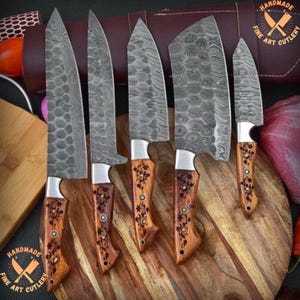 May include: A set of five kitchen knives with Damascus steel blades featuring distinct patterned textures. Each knife has a wooden handle decorated with small floral engravings and metal rivets. The blades vary in shape and size, including a cleaver.