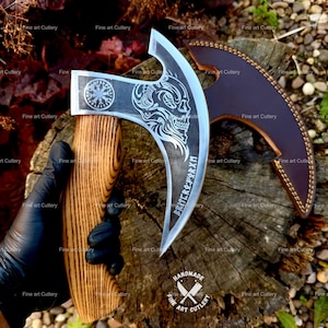 May include: A steel axe with a wooden handle featuring a skull engraving and runic text on the blade. The axe head displays a circular symbol and a detailed skull design. A dark brown leather sheath with stitched edges is positioned next to the axe.