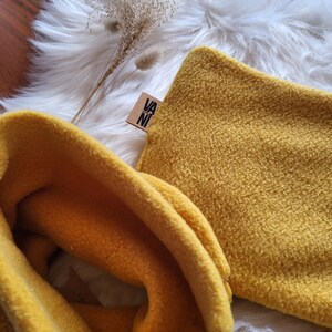 Könnte beinhalten: Zwei gelbe Fleece-Halswärmer in Senfgelb. Einer ist gefaltet, der andere gerollt. Der gefaltete Halswärmer hat ein kleines braunes Etikett mit dem Text "VANI" darauf.