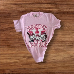 Może przedstawiać: Jasnor&oacute;żowa koszulka z grafiką przedstawiającą trzy postacie Hello Kitty grające w baseball. Tekst "HOUSTON ASTROS" jest wydrukowany nad i pod postaciami w czerwono-białym wzorze. Koszulka leży na drewnianej powierzchni.