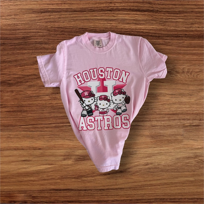 Może przedstawiać: Jasnor&oacute;żowa koszulka z grafiką przedstawiającą trzy postacie Hello Kitty grające w baseball. Tekst "HOUSTON ASTROS" jest wydrukowany nad i pod postaciami w czerwono-białym wzorze. Koszulka leży na drewnianej powierzchni.