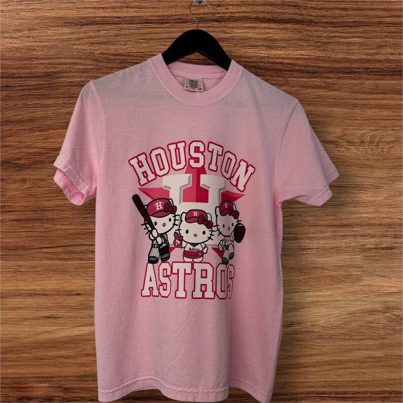 Może przedstawiać: Jasnor&oacute;żowa koszulka z napisem "HOUSTON ASTRO" w kolorze czerwono-białym i gwiazdą. Koszulka przedstawia trzy postacie Hello Kitty w strojach baseballowych, trzymające kij, piłkę i rękawicę.