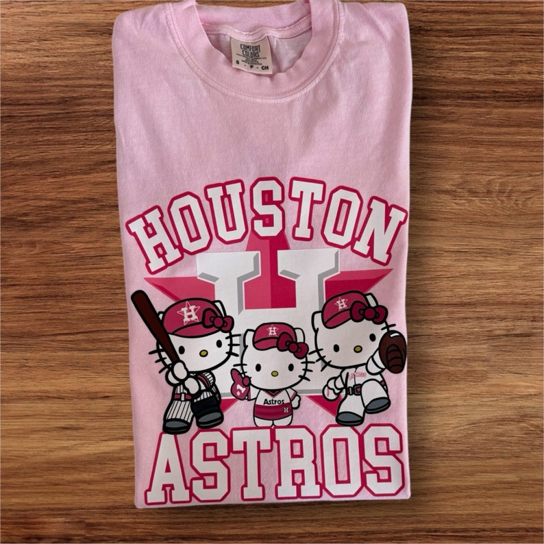 Może przedstawiać: Jasnor&oacute;żowa koszulka z napisem "Houston Astros" w kolorze białym i czerwonym. Koszulka przedstawia grafikę trzech postaci Hello Kitty w strojach baseballowych. Koszulka jest złożona na drewnianej powierzchni.