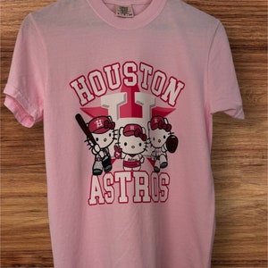 Może przedstawiać: Jasnor&oacute;żowa koszulka z napisem "HOUSTON ASTROS" w kolorze białym i czerwonym, z gwiazdą. Koszulka przedstawia r&oacute;wnież trzy postacie Hello Kitty w strojach baseballowych, trzymające kij, piłkę i rękawicę.