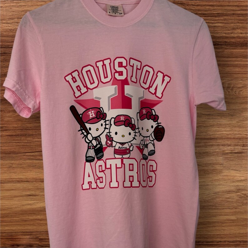 Może przedstawiać: Jasnor&oacute;żowa koszulka z napisem "HOUSTON ASTROS" w kolorze białym i czerwonym, z gwiazdą. Koszulka przedstawia r&oacute;wnież trzy postacie Hello Kitty w strojach baseballowych, trzymające kij, piłkę i rękawicę.