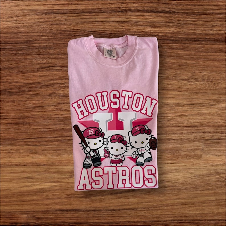 Może przedstawiać: Jasnor&oacute;żowa koszulka z napisem "Houston Astros" w kolorze białym i czerwonym. Koszulka przedstawia grafikę trzech postaci Hello Kitty w strojach baseballowych. Koszulka jest złożona na drewnianej powierzchni.